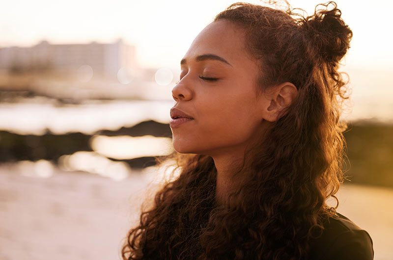 A woman stands at the beach with her eyes closed. It represents the calm that one can feel after hypnotherapy for anxiety relief.