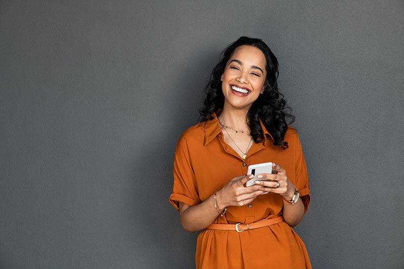 An image of a radiant, smiling woman standing confidently against a neutral gray wall. Her genuine joy reflects the positive impact of Career Fulfilment Coaching on her professional journey.